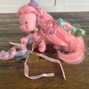 May include: A pink vintage toy figure with long pink hair and blue eyes. The figure has a gold collar and is adorned with small decorative elements. A pink ribbon and a gold charm are also present.