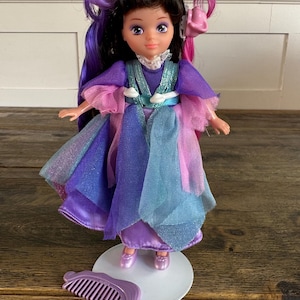 Op de afbeelding: Een pop met donkerbruin haar met paarse en roze lokken, gestyled met twee bloemclips. De pop draagt een gelaagde jurk in tinten paars, blauw en roze met een witte kanten kraag. Een paarse plastic kam ligt op de witte standaard bij de pop.