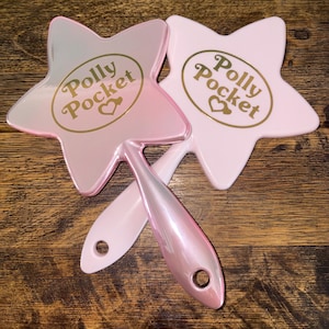 May include: Two star-shaped Polly Pocket hand mirrors. One is a light pink with a pearlescent handle, and the other is a solid light pink. The Polly Pocket logo is in gold lettering.