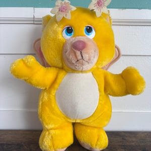 Könnte beinhalten: Ein Vintage-Teddybär aus gelbem Plüsch mit cremefarbenem Bauch und hellrosa Ohren. Der Bär hat blaue Augen, eine rosa Nase und zwei kleine Blumenakzente auf dem Kopf. Der Bär steht auf einer Holzoberfläche.