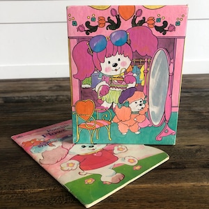 May include: A pink and green book with the title "Poochie Flowers" on the cover. The book features a cartoon illustration of a pink dog wearing sunglasses and a purple dress. The dog is standing in front of a mirror and a chair. The book is open to reveal a page with a cartoon illustration of a white dog wearing a red shirt and a pink heart.