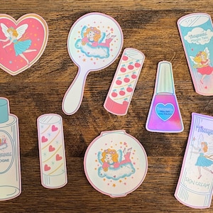 Puede incluir: Una colección de pegatinas con temática de hadas, incluyendo Campanilla. Las pegatinas representan artículos como un espejo, esmalte de uñas y otros cosméticos. La paleta de colores incluye rosa, azul y blanco, con detalles iridiscentes.