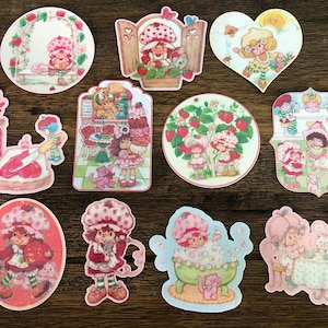 Peut inclure: Une collection d'autocollants colorés de Fraisinette de différentes formes. Les autocollants présentent le personnage dans différentes scènes, notamment un champ de fraises, un bain et une fête de thé. Les autocollants sont sur un fond en bois foncé.