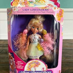 Könnte beinhalten: Lady Lovelylocks-Puppe in Originalverpackung. Die rosa Schachtel trägt die Aufschrift "Lady Lovelylocks" und "Enchanted Island". Die Puppe hat lange, blonde Haare mit rosa und gelben Akzenten und trägt ein Kleid.