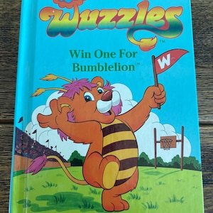 Könnte beinhalten: Ein Vintage-"Wuzzles"-Buch, "Win One For Bumblelion", zeigt einen Cartoon-Löwen, der eine Flagge mit einem "W" hält. Das Cover ist türkis mit farbenfrohem Text und Illustrationen. Collector Series Buch #4.