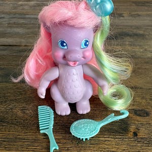 May include: A purple dragon doll with pink and light green hair, featuring a blue translucent frog accessory on its head. The doll has blue eyes and a textured chest. Included are a light blue plastic hair comb and a matching hair brush.