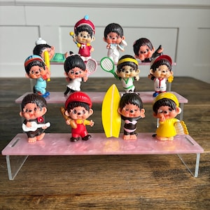 以下が含まれることがあります： さまざまなスポーツや活動をする漫画のキャラクターの小さなフィギュアのコレクション。フィギュアはピンクの段になったスタンドに展示されています。キャラクターは衣装を着て、スポーツ用品を持っています。