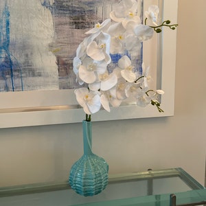 以下が含まれることがあります： 白い蘭の花が、質感のあるライトブルーのセラミックの花瓶に入っています。花瓶は、シルバーの金属フレームのガラスのテーブルの上に置かれています。