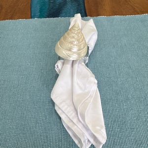 May include: A white cloth napkin held by a pearlescent seashell-shaped napkin ring. The napkin ring has a spiral design. The napkin is folded and placed on a light blue placemat.
