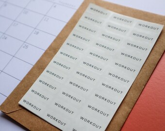 May include: A sheet of transparent stickers with the word "WORKOUT" printed in black, arranged in rows. The stickers are on a brown envelope, with a calendar and a coral-colored surface in the background. Ideal for fitness planning.
