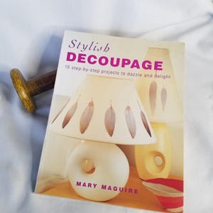 May include: A book titled "Stylish Decoupage" with a lamp illustration. The book cover features a white lamp with feather designs on the lampshade. A wooden object with a yellow handle is on the left.