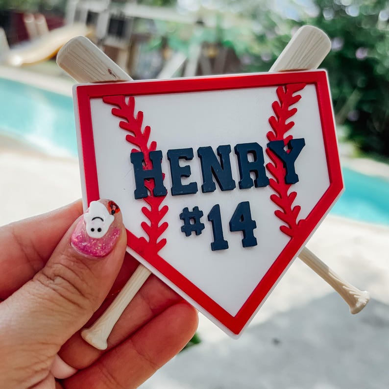 May include: A baseball-themed decoration featuring a white home plate design with a red border. The name "HENRY" and the number "#14" are displayed in navy blue. Two miniature baseball bats are crossed behind the home plate.