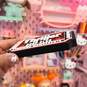 May include: A black, red, and white rectangular magnet with the text "Prayer Book Club" in a stylized font. The magnet is held in front of a pink pegboard with various decorative items.
