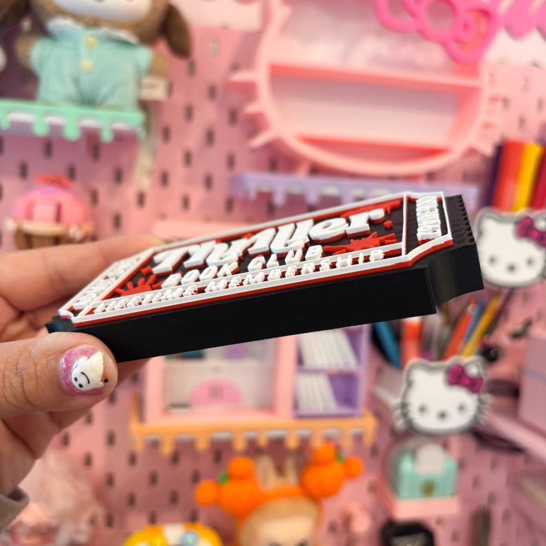 May include: A black, red, and white rectangular magnet with the text "Prayer Book Club" in a stylized font. The magnet is held in front of a pink pegboard with various decorative items.