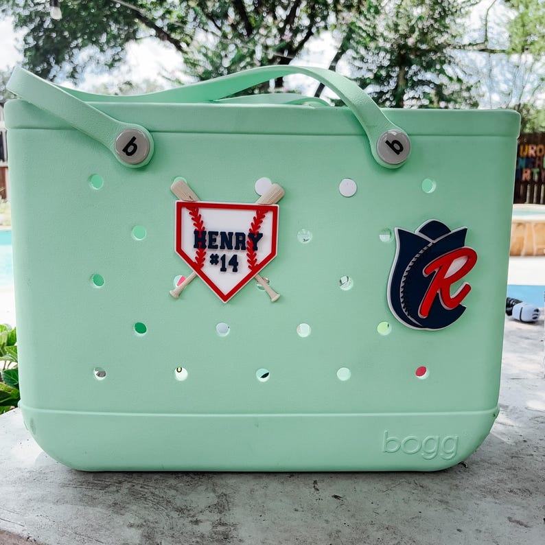 May include: A light green tote bag with a handle and the word "bogg" on the bottom. The bag has baseball-themed embellishments, including a baseball-shaped patch with the name "HENRY #14" and a baseball team logo.