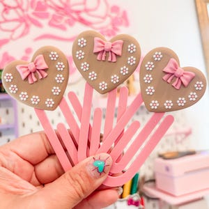 Puede incluir: Tres clips de papel rosas en forma de corazón con adornos marrones en forma de corazón. Cada adorno presenta un lazo rosa y detalles de flores blancas. Los clips de papel se sostienen en una mano, con un fondo rosa.