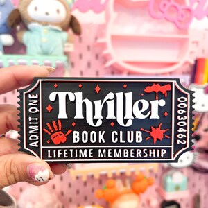 May include: A black and red "Thriller Book Club" ticket-shaped magnet. The magnet features white text, a red handprint, and blood splatter designs. The ticket reads "Lifetime Membership" and has a serial number.