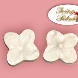 May include: Two white, flower-shaped fondant decorations on a pink background. The decorations have detailed petal designs and a slightly textured surface. The image includes the text "Icing Petals" in a gold script.