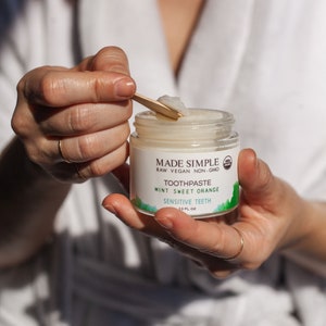 May include: A white jar of Made Simple Mint Sweet Orange Toothpaste for Sensitive Teeth. The jar is labeled with a green and white design and the text "RAW VEGAN NON-GMO".