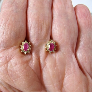 Puede incluir: Un par de pendientes de oro, cada uno con un rubí ovalado central rodeado de pequeñas piedras preciosas transparentes. Los pendientes se muestran sobre un fondo color piel, resaltando su diseño y el color del rubí.