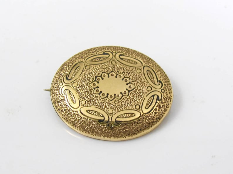 May include: A gold oval brooch with an intricate design of circles and swirls. The brooch has a textured surface and a pin on the back.