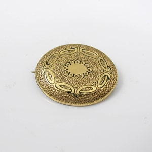 May include: A gold-toned brooch with a circular design. The brooch has a textured surface with a raised, circular pattern in the center.