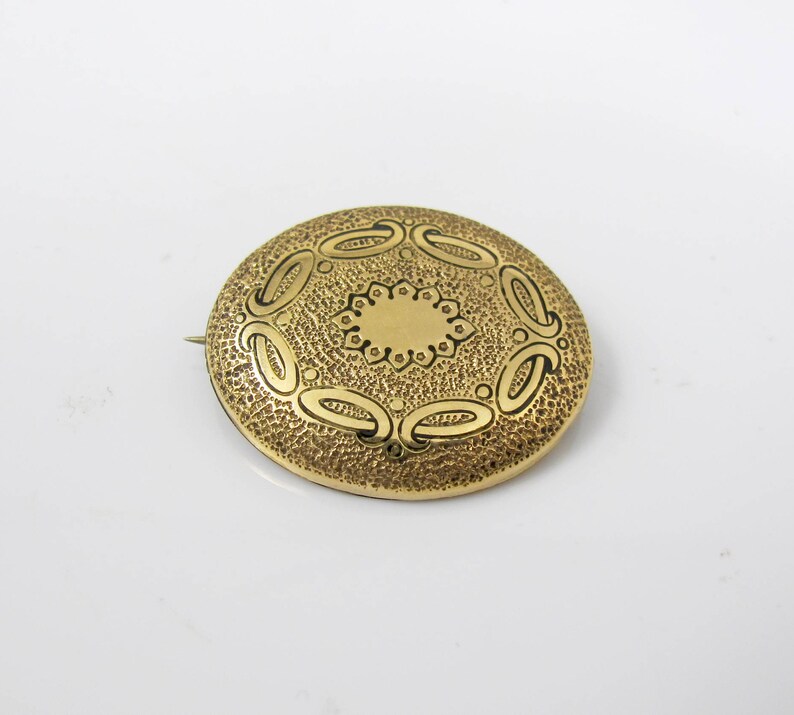 May include: A gold-toned brooch with a circular design. The brooch has a textured surface with a raised, circular pattern in the center.