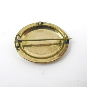 May include: The back of a gold oval brooch with a pin and catch. The brooch has a smooth, slightly textured surface.