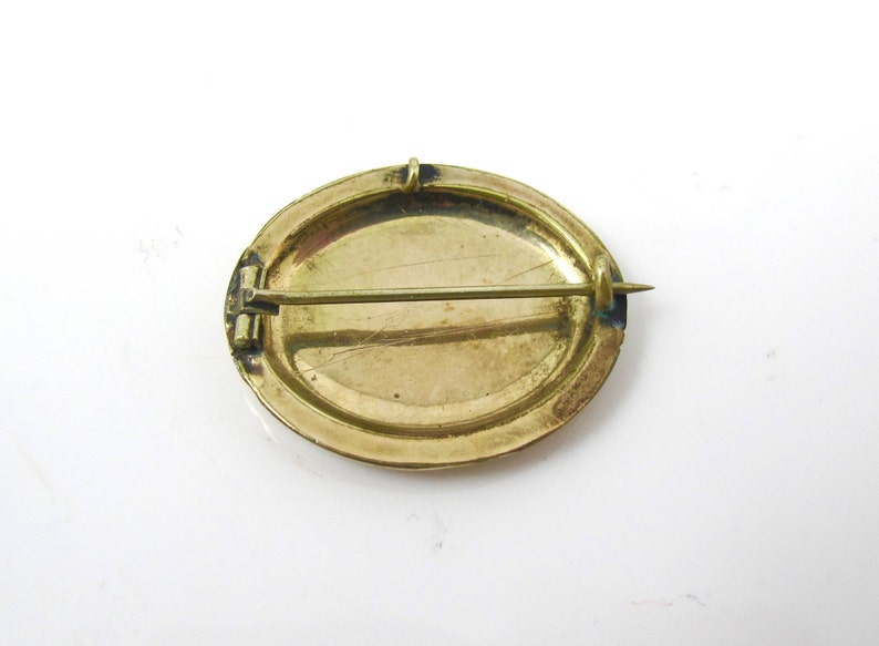 May include: The back of a gold oval brooch with a pin and catch. The brooch has a smooth, slightly textured surface.
