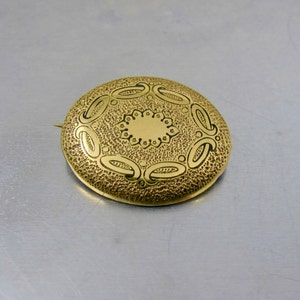May include: A gold-toned brooch with a circular design featuring a central circle surrounded by a ring of smaller circles and a ring of curved shapes.