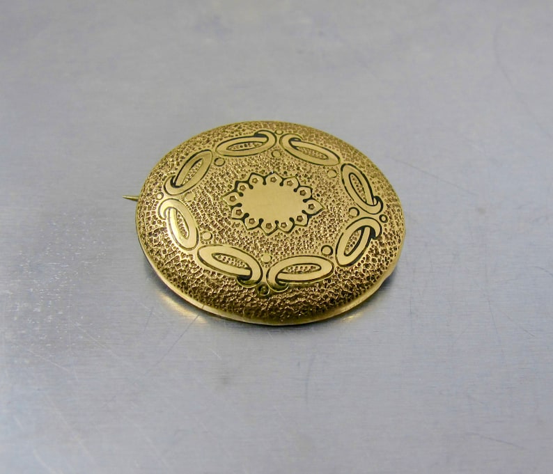 May include: A gold-toned brooch with a circular design featuring a central circle surrounded by a ring of smaller circles and a ring of curved shapes.