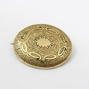 May include: A gold-toned brooch with a circular design. The brooch has a textured surface with a raised, circular pattern in the center.