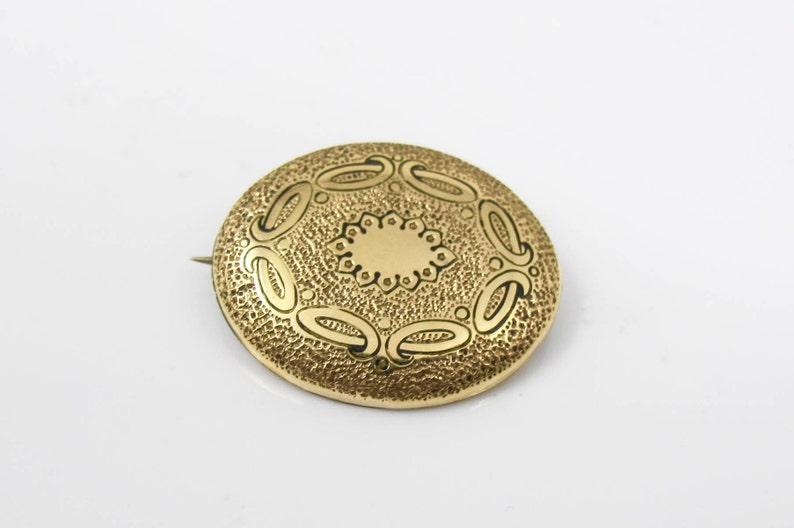 May include: A gold-toned brooch with a circular design. The brooch has a textured surface with a raised, circular pattern in the center.