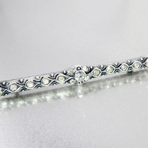 May include: A silver bar pin with a central clear rhinestone and smaller rhinestones along the sides. The pin has a decorative design with a black enamel finish.