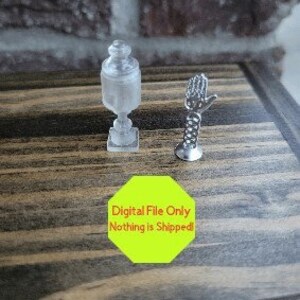 May include: Two small, clear glass figurines on a wooden surface. One is a decorative jar with a lid and the other is a hand with a decorative design.