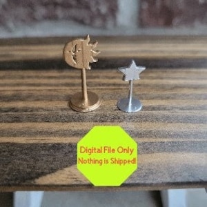 Könnte beinhalten: Zwei kleine dekorative Metallfiguren auf einer Holzoberfläche. Die größere Figur ist golden und zeigt ein Sonnen- und Mondmotiv. Die kleinere Figur ist silbern und zeigt ein Sternmotiv.