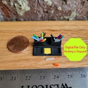 Può includere: Un organizzatore da scrivania nero con due scomparti per penne e pennarelli. L'organizzatore ha un piccolo blocco di note adesive giallo al centro. L'organizzatore è largo circa 7,6 cm e alto 5 cm.