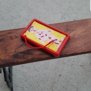 Puede incluir: Un marco de plástico rojo con fondo amarillo y una ilustración de dibujos animados de una persona con partes del cuerpo numeradas. El marco tiene una cuerda roja atada.