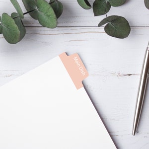 Puede incluir: Una hoja de papel blanca con una pestaña rosa etiquetada "Plato principal" sobre una superficie de madera blanca con un bolígrafo plateado y una rama de eucalipto.