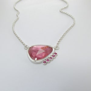 May include: A silver necklace featuring a pink gemstone pendant surrounded by smaller pink gemstones.