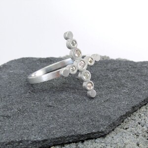 Può includere: Un anello d'argento con un design a forma di croce incastonato con piccoli diamanti trasparenti.