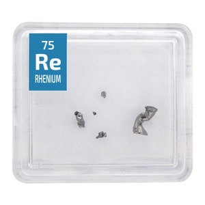 Może przedstawiać: Mały, przezroczysty pojemnik z plastiku z białą etykietą, na której widnieje napis "75 Re RHENIUM". Wewnątrz pojemnika znajduje się kilka małych kawałków srebrzystoszarego metalu.