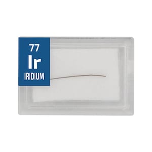 May include: A small sample of iridium, a hard, silvery-white metal, is displayed in a clear plastic case. The case has a blue label with the element's symbol "Ir" and atomic number "77". The label also reads "IRIDIUM".