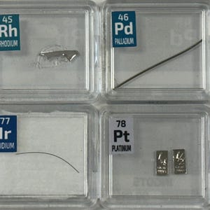 May include: A collection of eight small clear plastic containers, each containing a sample of a different element. The elements are labeled with their atomic number and symbol, and their names are written below. The elements are: Ruthenium, Rhodium, Palladium, Silver, Osmium, Iridium, Platinum, and Gold.