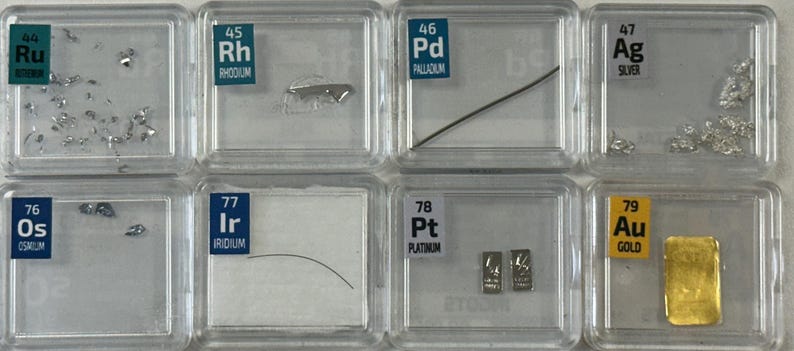 May include: A collection of eight small clear plastic containers, each containing a sample of a different element. The elements are labeled with their atomic number and symbol, and their names are written below. The elements are: Ruthenium, Rhodium, Palladium, Silver, Osmium, Iridium, Platinum, and Gold.