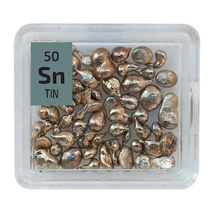 May include: A clear plastic container filled with small, silver-colored tin pellets. A label on the container reads "50 Sn TIN".