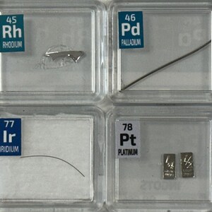 May include: A collection of eight clear plastic containers, each containing a sample of a different element. The elements are arranged in order of atomic number, from ruthenium (Ru) to gold (Au). Each container has a label with the element's name and atomic number.