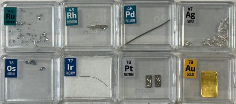 May include: A collection of eight clear plastic containers, each containing a sample of a different element. The elements are arranged in order of atomic number, from ruthenium (Ru) to gold (Au). Each container has a label with the element's name and atomic number.