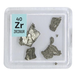 Puede incluir: Un pequeño contenedor de plástico con tapa transparente contiene varios trozos de circonio gris metálico, elemento 40. La etiqueta dice "40 Zr ZIRCONIUM".