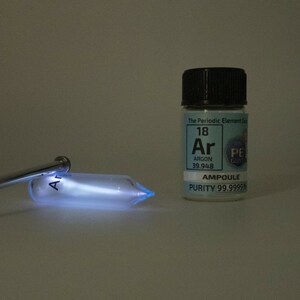 Puede incluir: Un tubo de vidrio lleno de gas argón brilla en azul cuando se hace pasar una corriente eléctrica a través de él. Una pequeña botella etiquetada "Argón 18 Ar 39.948 AMPOULE PUREZA 99.9999%" se encuentra junto al tubo.